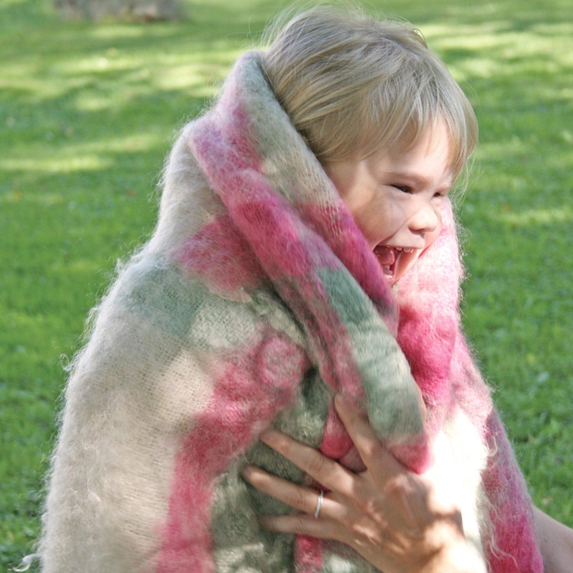 A young child with blond hair is wrapped in a Victorian Bouclé Spring Garden Woven Blanket featuring colorful, fluffy patterns of pink and green. The child laughs joyfully as a hand with a ring gently holds the blanket close. The background displays a grassy area bathed in sunlight, evoking the charm of Halcyon Yarn's creation.