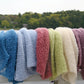 A row of vibrantly colored Victorian Bouclé Stoles from Halcyon Yarn draped over a white wooden railing. The stoles come in blue, light blue, red, green, cream, and purple colors. A serene lake and tree-lined shore are visible in the background under a cloudy sky.