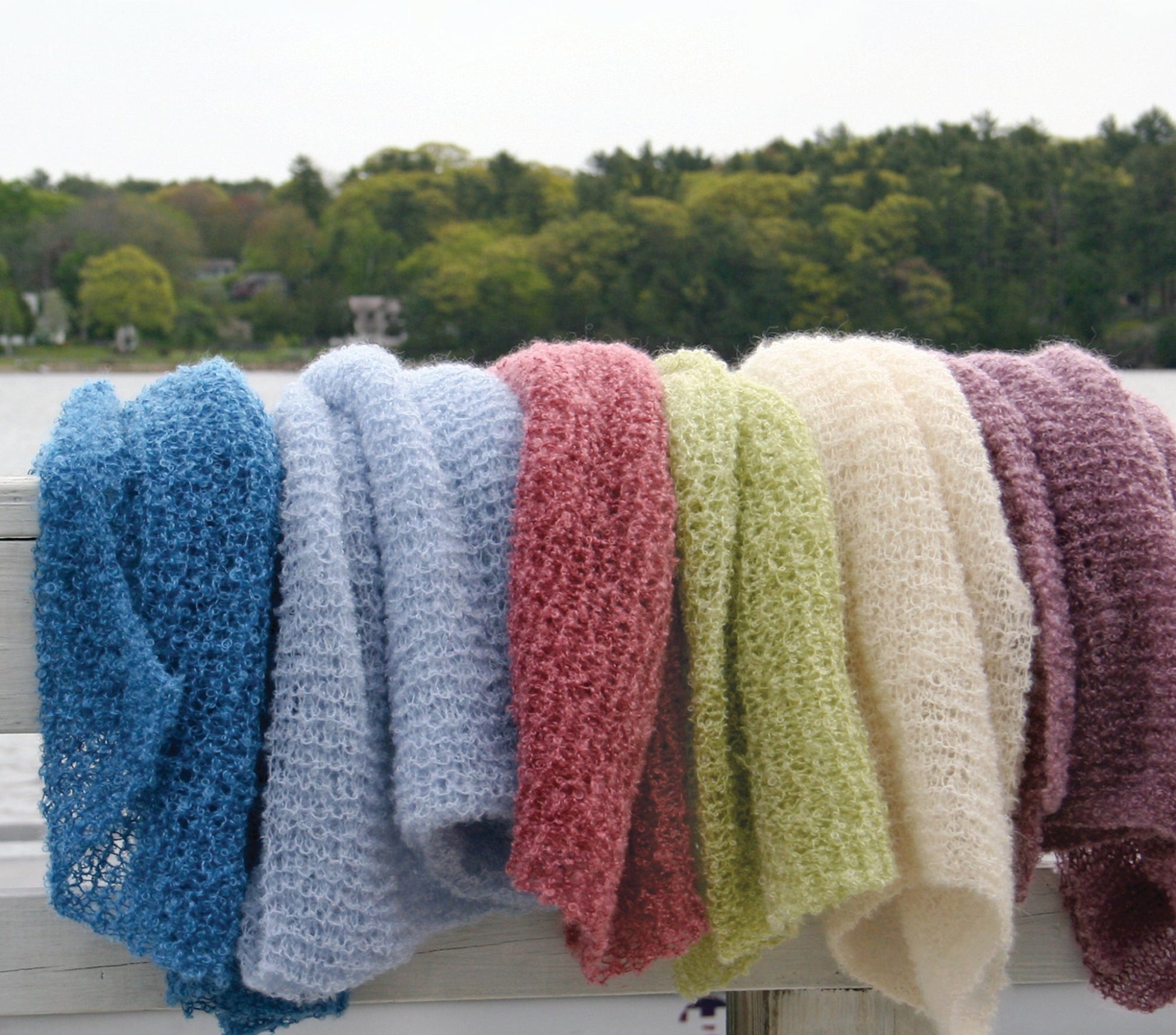 A row of vibrantly colored Victorian Bouclé Stoles from Halcyon Yarn draped over a white wooden railing. The stoles come in blue, light blue, red, green, cream, and purple colors. A serene lake and tree-lined shore are visible in the background under a cloudy sky.