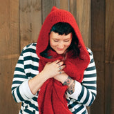 A person with short black hair, a tattoo on their right hand and left forearm is dressed in a black and white striped shirt and a red hooded Gimme Shelter scarf from Halcyon Yarn that resembles Victorian Bouclé. They stand in front of a wooden wall, looking down with a gentle smile, holding the scarf close as if savoring cozy winter days.