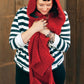 A person with short dark hair and bangs is wearing a black-and-white striped sweater, black skirt, and a red Gimme Shelter hooded scarf with pockets by Halcyon Yarn. They are standing against a wooden background, looking down and holding the scarf with both hands, appearing cozy and content on winter days.