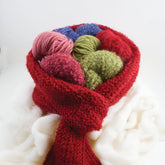 The "Sophisticated Weekend Scarf" from Halcyon Yarn cradles several balls of colorful yarn, including pink, green, blue, and red. The setup rests on a soft, white, fluffy surface, evoking a touch of Victorian Boucle charm.