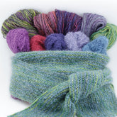 The Sophisticated Simplicity Scarf by Halcyon Yarn, a soft knitted accessory in a greenish-blue hue with a textured pattern, is shown tied in a knot. Inside the scarf are balls and skeins of colorful Lana Bambu yarn in vibrant shades of purple, red, pink, light blue, and green. The variety of yarn materials adds visual interest to this delightful display.