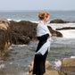 A woman with light hair pulled into a bun is standing on a rocky shoreline, looking out over the ocean. She is wearing a long black dress and an elegant Victorian Bouclé Crochet Stole by Halcyon Yarn draped over her shoulders. White waves crash against the rocks, and the sky is clear.