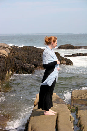 A woman with light hair pulled into a bun is standing on a rocky shoreline, looking out over the ocean. She is wearing a long black dress and an elegant Victorian Bouclé Crochet Stole by Halcyon Yarn draped over her shoulders. White waves crash against the rocks, and the sky is clear.