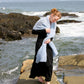 A woman adorned in a long black dress and the luxurious Victorian Bouclé Crochet Stole from Halcyon Yarn strolls barefoot on rocks by the ocean. Waves crash against the rugged coastline behind her as she gazes downward, her hair elegantly tied up. The sky is clear, painting a serene and peaceful backdrop.