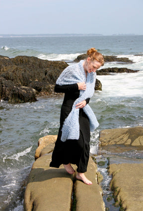 A woman adorned in a long black dress and the luxurious Victorian Bouclé Crochet Stole from Halcyon Yarn strolls barefoot on rocks by the ocean. Waves crash against the rugged coastline behind her as she gazes downward, her hair elegantly tied up. The sky is clear, painting a serene and peaceful backdrop.