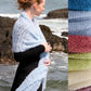 A person wearing a black top stands by the rocky seaside, wrapped in an elegant Victorian Bouclé Crochet Stole from Halcyon Yarn in light blue. A side panel displays various colors of similar crochet pattern fabric: light blue, white, pink, red, yellow, and green. Waves crash gently against the rocks.