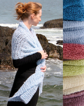 A person wearing a black top stands by the rocky seaside, wrapped in an elegant Victorian Bouclé Crochet Stole from Halcyon Yarn in light blue. A side panel displays various colors of similar crochet pattern fabric: light blue, white, pink, red, yellow, and green. Waves crash gently against the rocks.