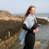 A woman stands on a rocky shoreline, wearing a light blue knit shawl over a black dress. She gazes out towards the ocean with her hair gently blowing in the breeze. The sky is clear and the sea is calm, with waves lightly crashing against the rocks; an elegant Victorian Bouclé Crochet Stole by Halcyon Yarn completes her serene look.