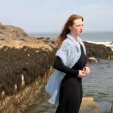 A woman with long, light brown hair stands on rocky, seaweed-covered terrain by the ocean. She is wearing a black dress and an elegant Victorian Bouclé Crochet Stole from Halcyon Yarn, gazing off into the distance with a serene expression. The sky above is partly cloudy.