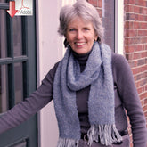 A woman with short gray hair, wearing a gray turtleneck and a long, blue Halcyon Yarn Frosted Elegance Scarf, smiles as she stands beside a door. The background shows a brick wall and part of a window. An Adobe logo with a downward arrow is displayed in the top left corner indicating "download.