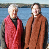 Two women are standing outdoors near a body of water with a tree-lined shore in the background. The older woman on the left is wearing a Halcyon Yarn Victorian Bouclé Wrap in red, and the younger woman on the right is wearing a brown Victorian Bouclé Wrap along with a stylish vest. Both are smiling at the camera.