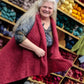 A woman with long white hair smiles while standing in front of shelves filled with colorful yarns. She is wearing a patterned top and an oversized, textured red vest, which she holds out to the sides, creating movement in the fabric. Her stylish Victorian Bouclé Wrap from Halcyon Yarn hints at an easy knitting pattern for enthusiasts.