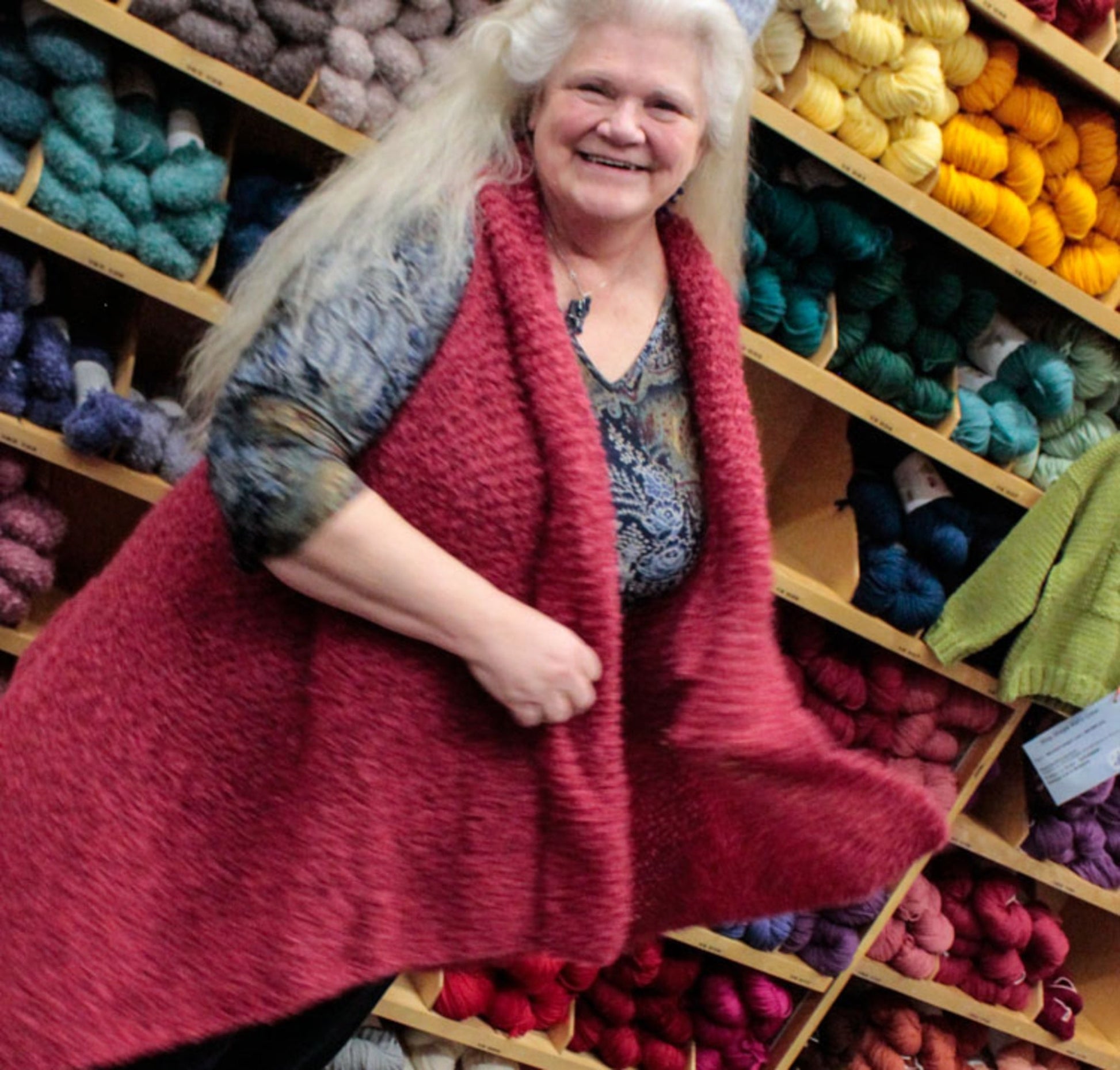 A woman with long white hair smiles while standing in front of shelves filled with colorful yarns. She is wearing a patterned top and an oversized, textured red vest, which she holds out to the sides, creating movement in the fabric. Her stylish Victorian Bouclé Wrap from Halcyon Yarn hints at an easy knitting pattern for enthusiasts.