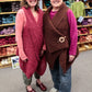 Two women are standing in a yarn store, smiling at the camera. They both wear stylish vests; the woman on the left has a red vest, and the woman on the right has a brown vest with a wooden clasp. Shelves filled with colorful yarn are visible in the background, including an easy knitting pattern for Halcyon Yarn's downloadable Victorian Bouclé Wrap.