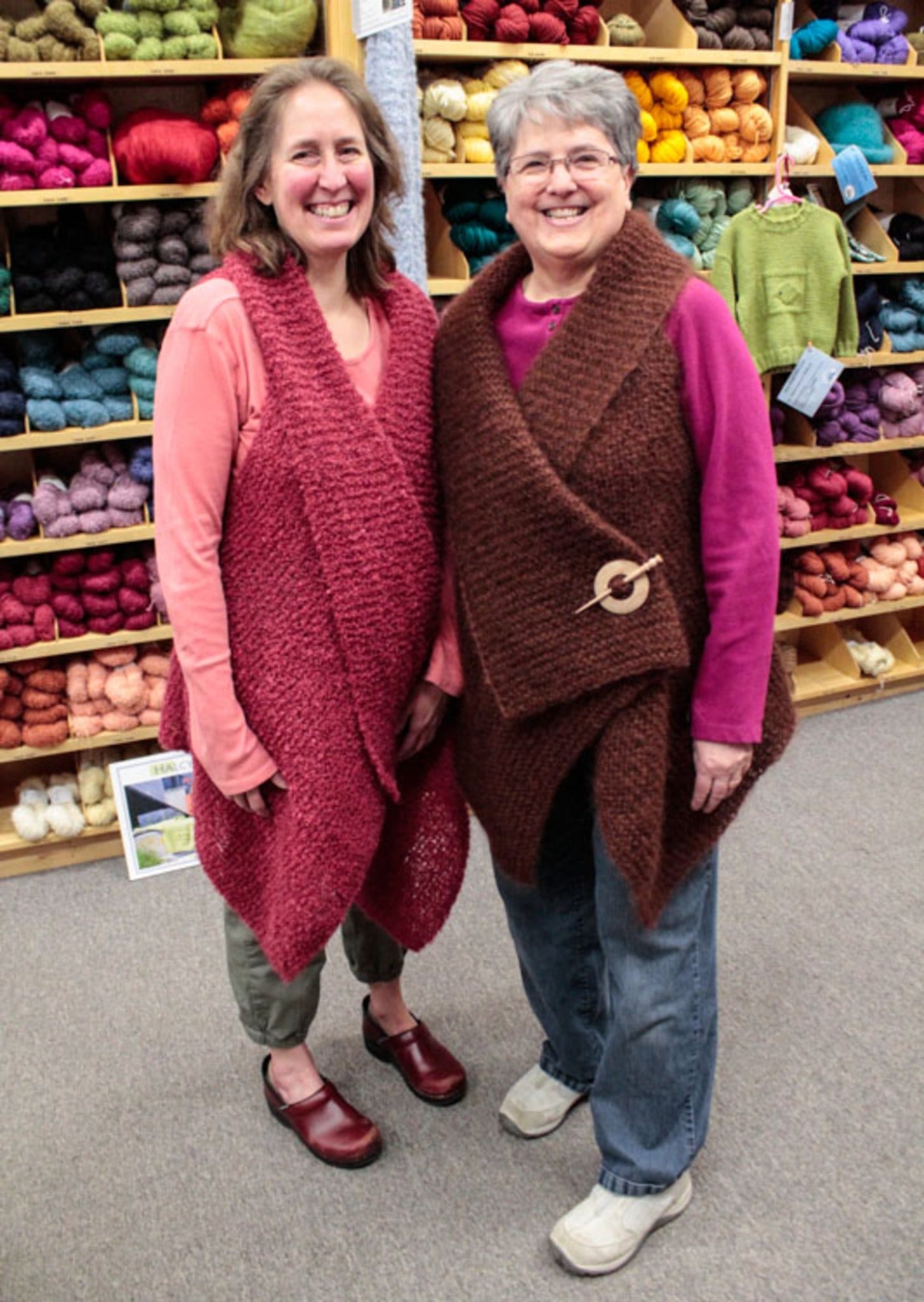 Two women are standing in a yarn store, smiling at the camera. They both wear stylish vests; the woman on the left has a red vest, and the woman on the right has a brown vest with a wooden clasp. Shelves filled with colorful yarn are visible in the background, including an easy knitting pattern for Halcyon Yarn's downloadable Victorian Bouclé Wrap.