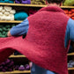 A person wearing a pink handmade sweater, facing away, is enveloped in layers of thick yarn reminiscent of a Victorian Bouclé Wrap by Halcyon Yarn. They stand before a wooden shelf filled with various colorful skeins of yarn in shades of green, blue, purple, orange, and white.