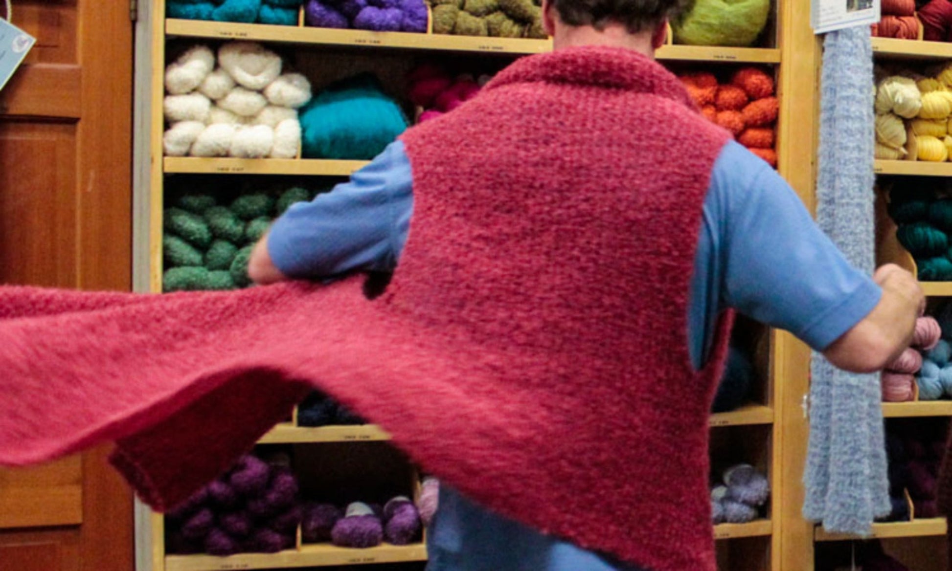 A person wearing a pink handmade sweater, facing away, is enveloped in layers of thick yarn reminiscent of a Victorian Bouclé Wrap by Halcyon Yarn. They stand before a wooden shelf filled with various colorful skeins of yarn in shades of green, blue, purple, orange, and white.