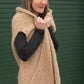 A woman with shoulder-length brown hair smiles while standing in front of a green door. She is wearing a beige Bouclé Cocoon Vest from Halcyon Yarn, which is sleeveless and hooded, over a long-sleeved dark shirt. She holds the front of her cozy vest with both hands.