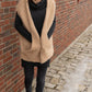 A woman with shoulder-length brown hair stands on a cobblestone pathway next to a brick wall. She is wearing a black outfit with a cozy beige Bouclé Cocoon Vest by Halcyon Yarn and brown ankle boots, with her hands in her pockets. The ground is partially covered with snow and ice.