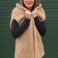 A woman with a fringe is wearing a tan-colored Bouclé Cocoon Vest from Halcyon Yarn over a dark sweater and pants. She is standing against a green wooden background and holding the vest closed at the chest with a shy smile.