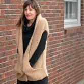 A woman with medium-length brown hair stands against a brick wall, wearing a black long-sleeve top and the Bouclé Cocoon Vest by Halcyon Yarn, expertly crafted using modular knitting techniques. She smiles softly and looks slightly to the side. A window is visible in the brick wall behind her.