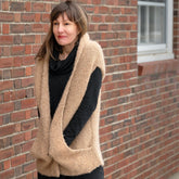 A woman with shoulder-length brown hair stands against a brick wall, wearing a cozy, light brown Bouclé Cocoon Vest from Halcyon Yarn over a black outfit. Her hands are tucked into the open sides of the vest, and she has a slight smile on her face.