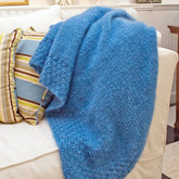 A luxurious, Lush Victorian Mohair Throw from Halcyon Yarn, in a cozy blue hue, is draped over the arm of a white sofa. The sofa features green, blue, and white striped pillows that add a splash of color to the neutral backdrop of the room. The scene evokes a sense of comfort and invitation in this living space.