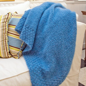 A cozy, blue Lush Victorian Mohair Throw from Halcyon Yarn is draped over a white sofa. The throw appears soft and textured, adding a pop of color to the setting. Behind it are several striped and patterned pillows in shades of blue, green, yellow, and white.