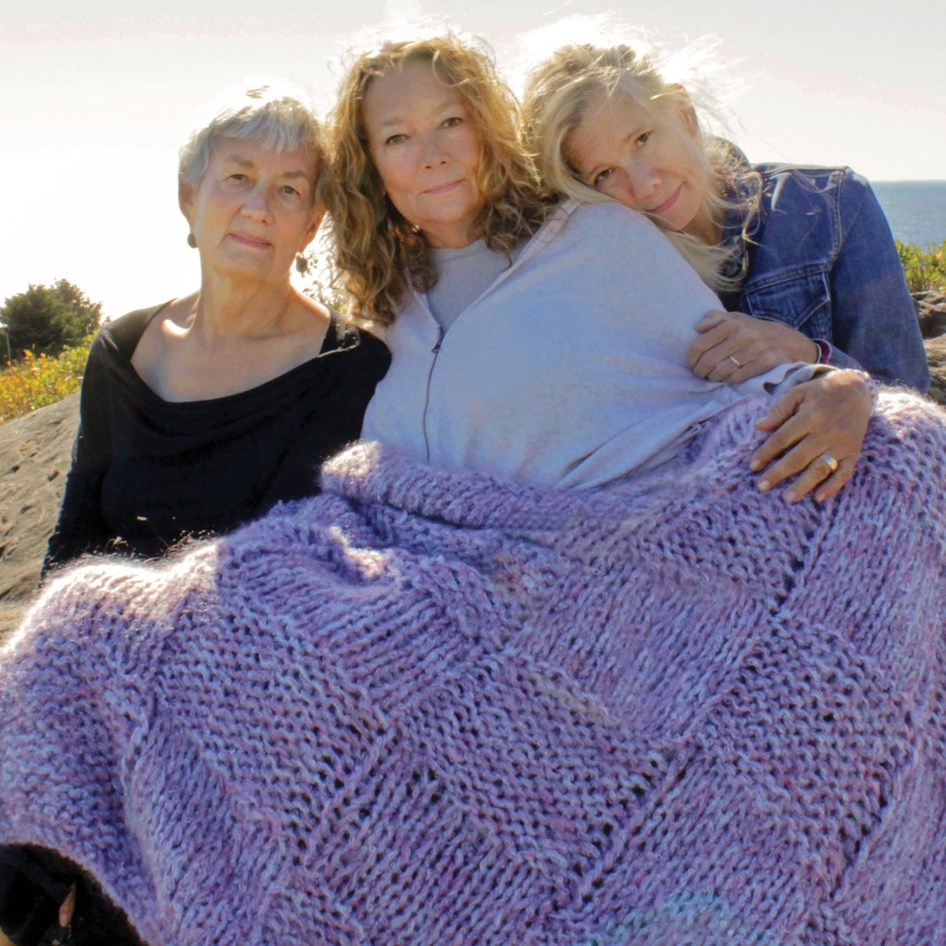 Three women sit closely together outdoors, with the ocean in the background. The woman on the left has short gray hair and wears a black top, the middle woman has curly light brown hair and wears a light top, and the third woman leans on her, all wrapped in Halcyon Yarn’s luxurious Cobble Stone Cove Throw.