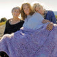 Three women sit close together on a rock with a scenic background, wrapped in a large, chunky knit purple Cobble Stone Cove Throw from Halcyon Yarn. The woman on the left has short gray hair, the middle woman has curly hair, and the woman on the right has long, light hair.