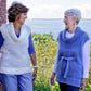 Two women are smiling and having a conversation outdoors beside a brick wall. The woman on the left is wearing jeans and a white sleeveless sweater reminiscent of Victorian style. The woman on the right sports Halcyon Yarn's Seguin Coveside Tunic with a belt and light blue pants. A scenic body of water is in the background, adding to the Halcyon Yarn-like tranquility.