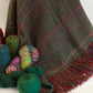 A richly woven Halcyon Yarn Forest Glen Broken Twill Throw, featuring red and multicolored threads, hangs elegantly, showcasing its fringed edge. Surrounding the blanket are skeins and cones of super bulky accent yarn in vibrant shades of red, green, blue, and multicolor, enhancing the cozy and colorful textures.