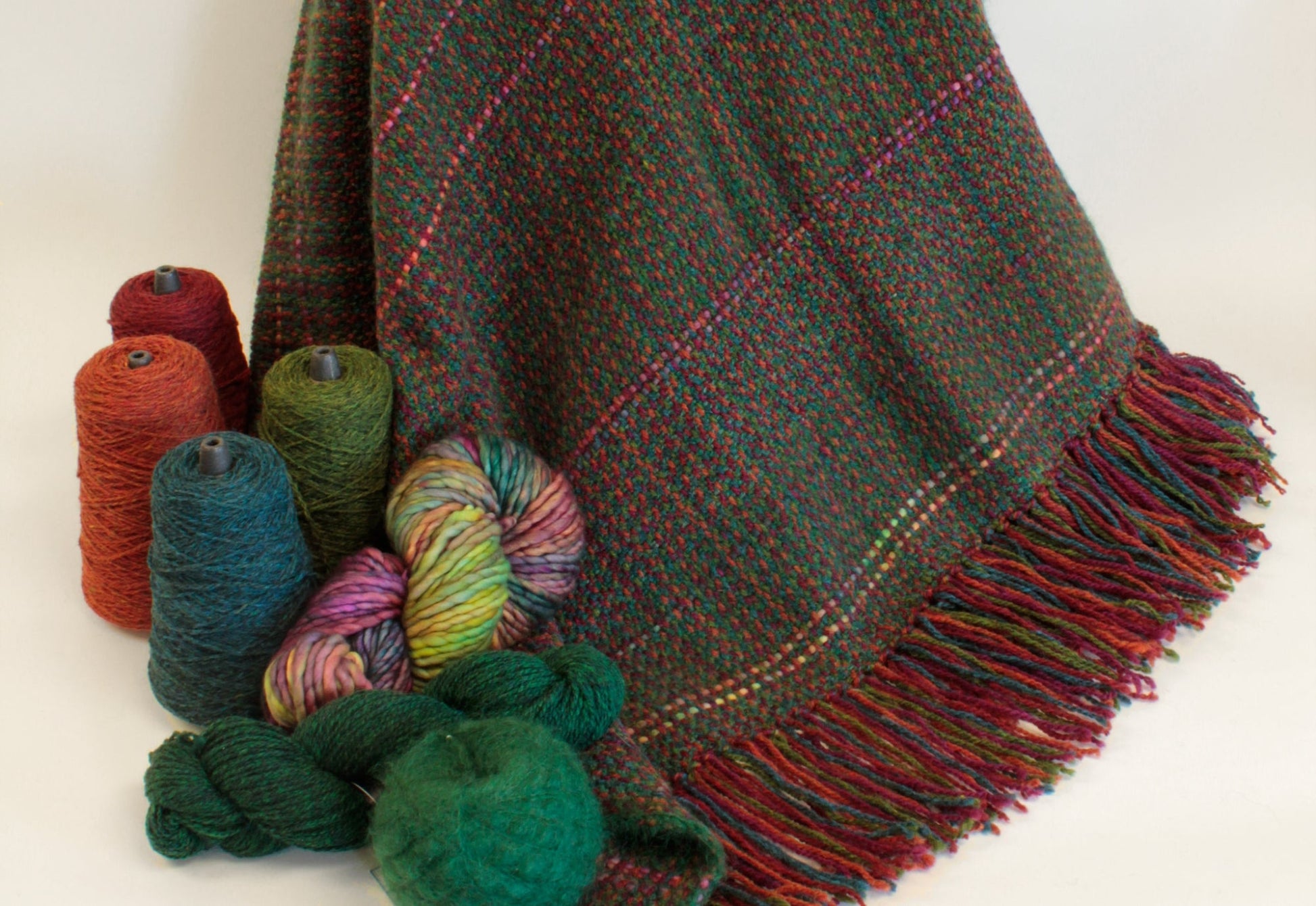 A richly woven Halcyon Yarn Forest Glen Broken Twill Throw, featuring red and multicolored threads, hangs elegantly, showcasing its fringed edge. Surrounding the blanket are skeins and cones of super bulky accent yarn in vibrant shades of red, green, blue, and multicolor, enhancing the cozy and colorful textures.