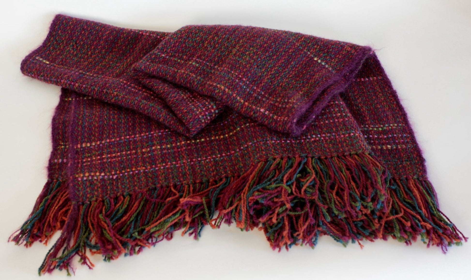 Introducing the Forest Glen Broken Twill Throw by Halcyon Yarn—a folded, multicolored wool scarf adorned with a plaid pattern and fringe ends. This exquisite scarf features an intricately woven broken twill design in shades of red, purple, and green. The harmonious blend of colors and detailed craftsmanship is beautifully showcased against a plain white background.