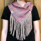 A person wearing a black shirt models the Obladi Oblada Woven Fringed Cowl by Halcyon Yarn, which has a unique triangular shape. This scarf is woven on a rigid heddle loom and showcases light purple and pink hues, secured with two buttons. Long beige fringe adorns the bottom edge, set against a wooden background.