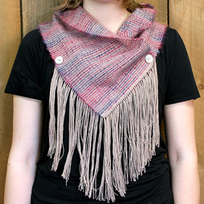 A person wearing a black shirt models the Obladi Oblada Woven Fringed Cowl by Halcyon Yarn, which has a unique triangular shape. This scarf is woven on a rigid heddle loom and showcases light purple and pink hues, secured with two buttons. Long beige fringe adorns the bottom edge, set against a wooden background.