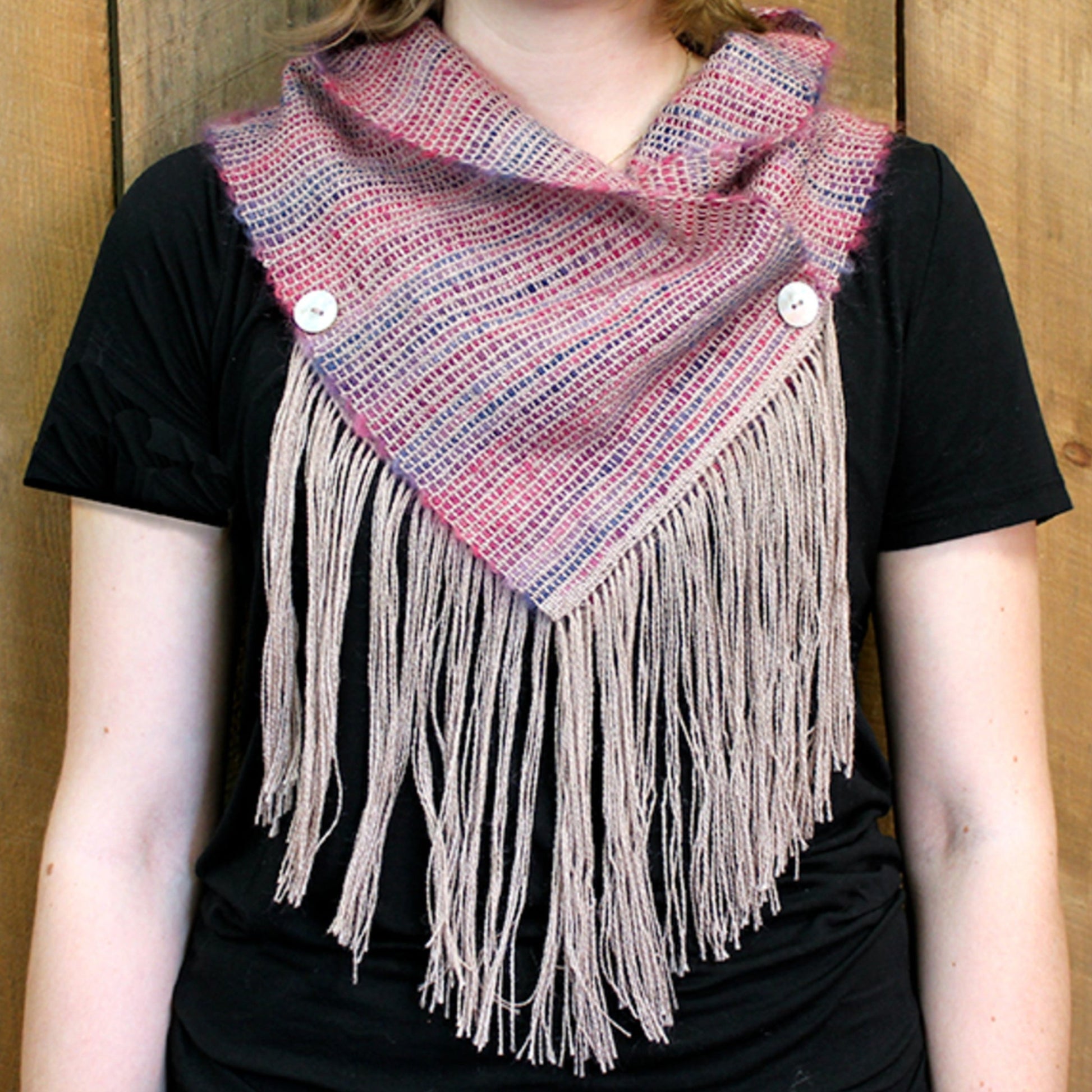 A person is wearing the Obladi Oblada Woven Fringed Cowl by Halcyon Yarn, an airy and lightweight cowl in shades of pink and white. The cowl features a triangular shape with long fringes along the bottom edge and is fastened with two white buttons at the shoulders. The person has paired it with a black shirt.