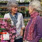 Two elderly women with gray hair are engaged in conversation in a garden center. Both are wearing "Oh So Sari Scarves!" from Halcyon Yarn; one blue and white, the other pink and red. They are surrounded by lush greenery and blooming pink flowers. A PDF icon indicating the scarf download is visible in the bottom left corner.