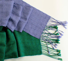 A close-up of the Shimmering Shadow Weave Scarf by Halcyon Yarn, showcasing its two-toned design with one side in lavender and the other in green. Crafted from Tencel yarn, the lavender side displays a herringbone pattern and tasseled fringes, while the green side features a more uniform texture. Spread out on a white background, this scarf is perfect for weaving projects.