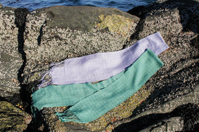 Two Shimmering Shadow Weave Scarves from Halcyon Yarn, one in light purple and the other in green, are draped across barnacle-covered rocks near the edge of the water. The texture and details of these Tencel yarn creations are highlighted by the natural light.