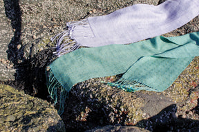 Two Shimmering Shadow Weave Scarves from Halcyon Yarn, one in lavender and one in teal, are draped over rocks encrusted with barnacles and seaweed. Featuring fringe at the ends, these scarves made from Tencel yarn contrast beautifully with the rough textures of the rocks. The scene is brightly lit by sunlight.