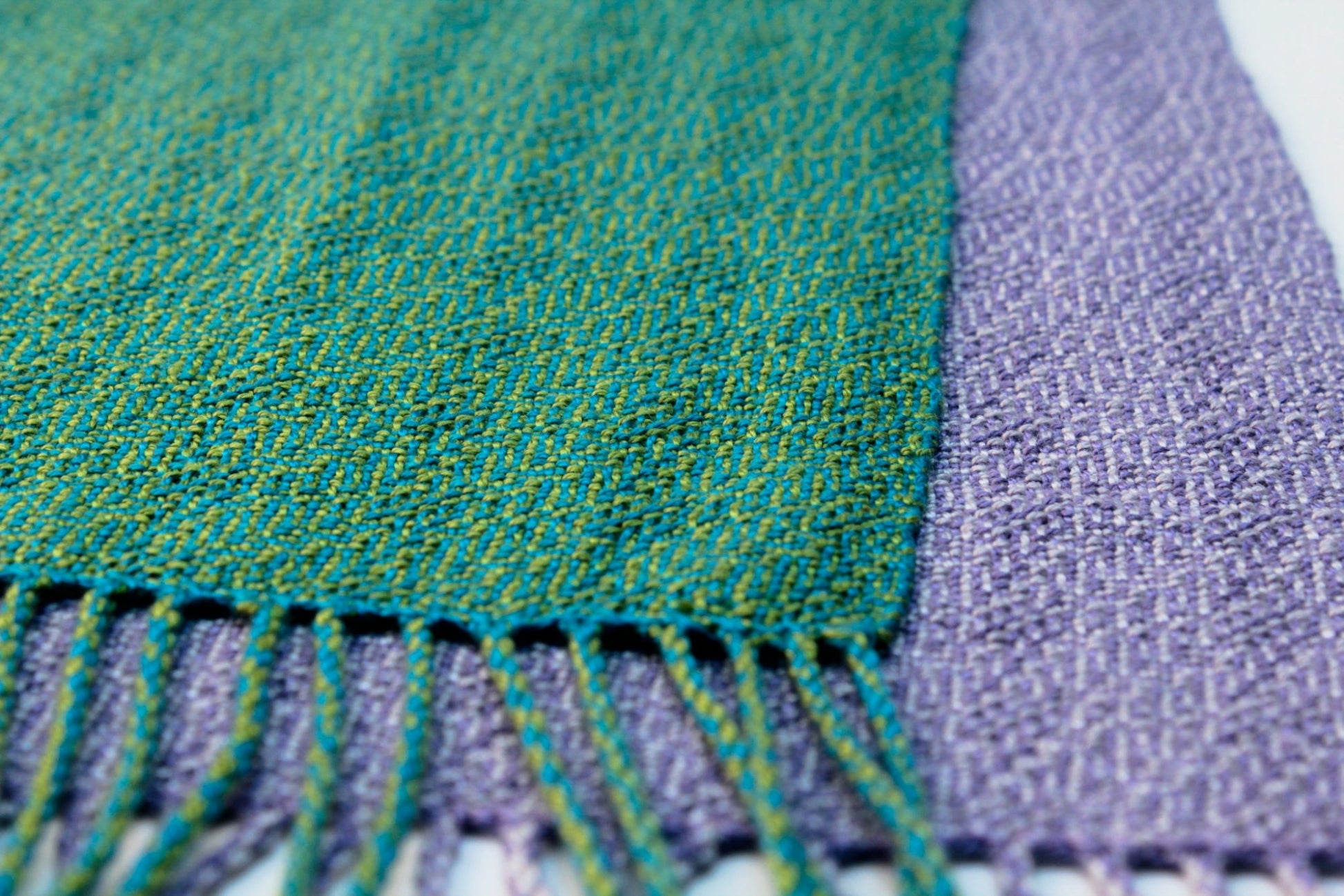 A close-up image showcases two handwoven fabric pieces from recent weaving projects. The piece on the left, crafted from Tencel yarn, boasts a green hue interspersed with blue and yellow threads. On the right is a lilac Halcyon Yarn Shimmering Shadow Weave Scarf, downloadable. Both fabrics are adorned with fringed edges and exhibit a soft yet textured appearance.