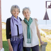 Two older women, both with short, grey hair, stand arm in arm and smiling outside on a sunny day. Both are wearing Halcyon Yarn's Shimmering Shadow Weave Scarves—one in lavender and the other in green. Behind them is an ocean view with clear skies and part of a brick building.