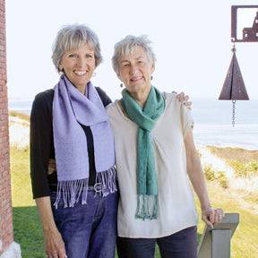 Two elderly women stand close together, smiling at the camera. One wears a purple Shimmering Shadow Weave Scarf, and the other wears a green one. They stand outdoors near a brick wall, with a scenic ocean view and grassy landscape in the background, proudly displaying their latest weaving projects from Halcyon Yarn.