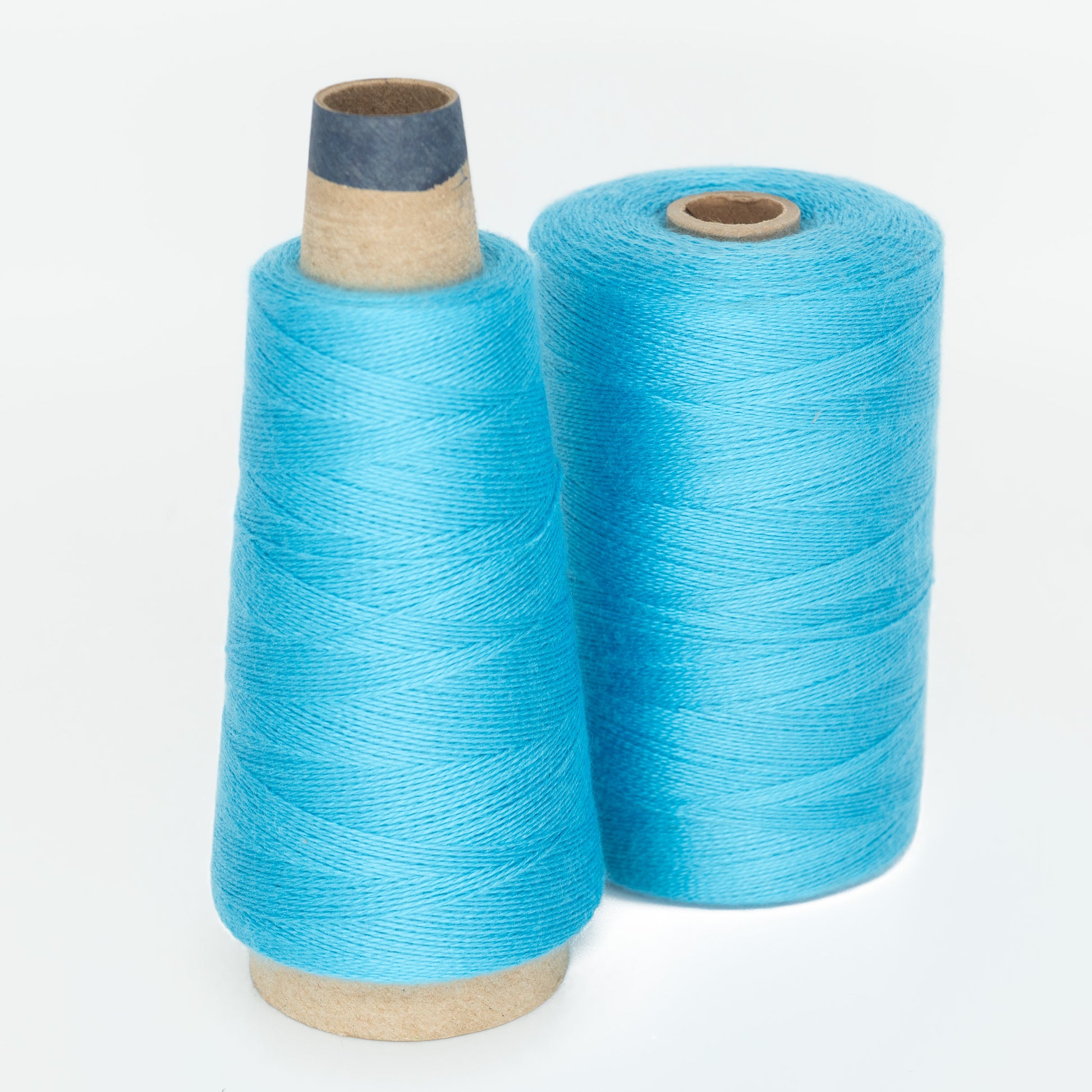 Two spools of vibrant 8/2 Tencel Yarn from Maurice Brassard, perfect for weaving projects, are pictured against a white background. The spool on the left is shaped like a cone, while the spool on the right is cylindrical. Both spools feature a similar shade of neatly wound blue thread.