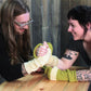 Two people arm wrestle across a wooden table, both wearing Halcyon Yarn's Whole Wide World Fingerless Mitts in oversized, color-blocked yellow and white wool. The person on the left has long hair and glasses, while the person on the right has short hair and visible arm tattoos. They are both smiling brightly, enjoying the moment.