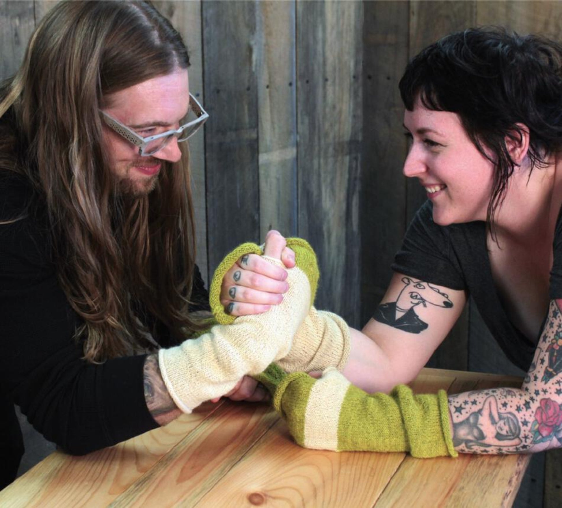 Two people arm wrestle across a wooden table, both wearing Halcyon Yarn's Whole Wide World Fingerless Mitts in oversized, color-blocked yellow and white wool. The person on the left has long hair and glasses, while the person on the right has short hair and visible arm tattoos. They are both smiling brightly, enjoying the moment.