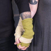 A close-up photo of two people holding hands. One person is wearing a black long-sleeved shirt with a tattoo visible on their forearm, while the other wears a grey shirt. Both are sporting Halcyon Yarn's Whole Wide World, Fingerless Mitts in beige and green-beige stripes—perfect for cozy days.